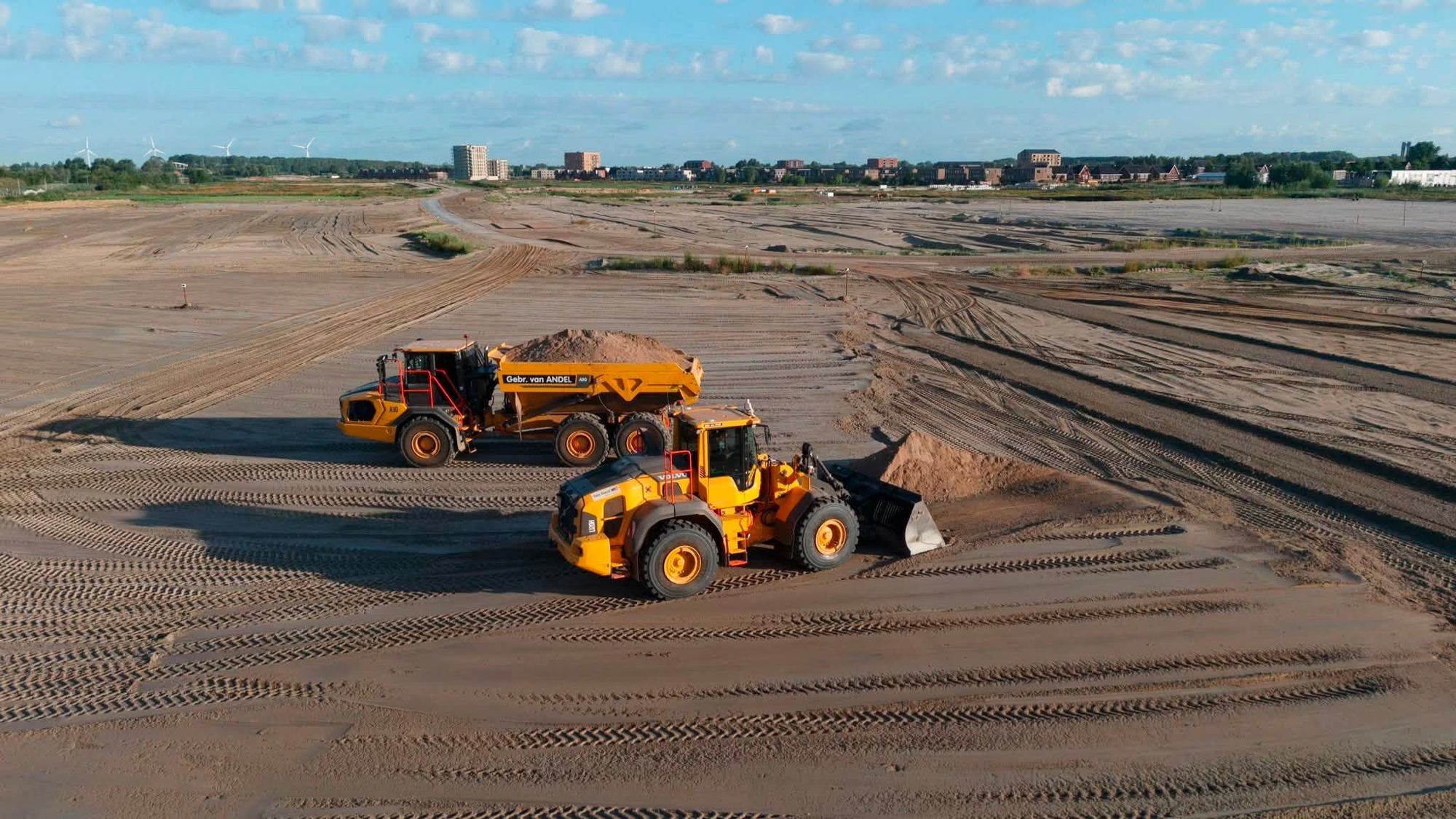 Knikdumper en wiellader naast elkaar met nieuwbouw op de achtergrond